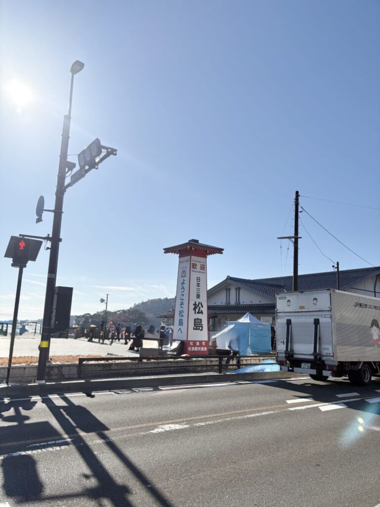 「青空の下、松島駅前の大きな看板（“ようこそ松島へ”）と、道路に停まるトラック、歩道を歩く観光客が写っている松島の風景。遠くに海や小島が見え、観光地のにぎわいが感じられる写真。」