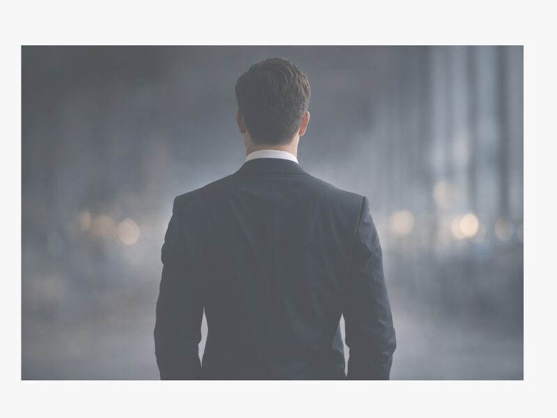スーツを着た男性の後ろ姿。ぼかした背景の中で立っているシンプルな構図。