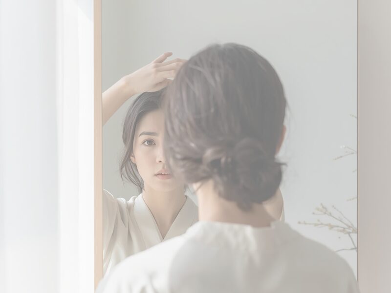 鏡を見ながら頭頂部の分け目を気にする女性の様子（女性の薄毛・てっぺんが気になるイメージ）